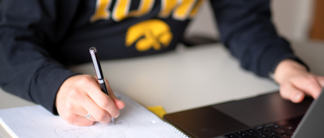 student writing in a notebook with a laptop in front of them